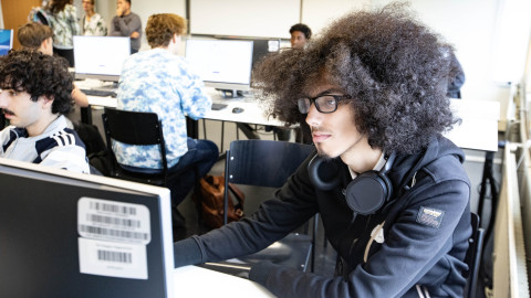 Een jongeman zit in een leslokaal achter een computer waarop hij aan het werk is. Om hem heen zitten andere studenten achter de computer.