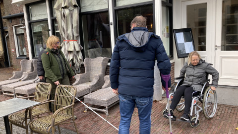 Een foto in de winkelstraat op Vlieland waar een persoon met witte stok loopt, een persoon in een rolstoel rijdt en een begeleider te zien is