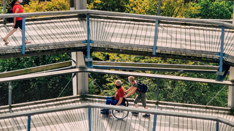 De foto bevat twee loopbruggen boven elkaar waarbij op een van de loopbruggen iemand in een rolstoel wordt voortgeduwd