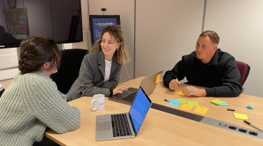 Drie mensen zitten met laptops aan tafel in een kantoorruimte. Ze zijn in gesprek en lachen. Op tafel liggen posts-its.