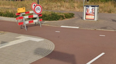 Een onveilige en onoverzichtelijke verkeerssituatie voor voetgangers met obstakels zoals een verkeersbord op het fietspad en een elektriciteitskast op de stoep aan het einde van de geleidelijnen.